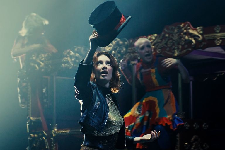 Stage performer holding a black top hat in a musical production inspired by The Greatest Showman, with performers on stage in background.